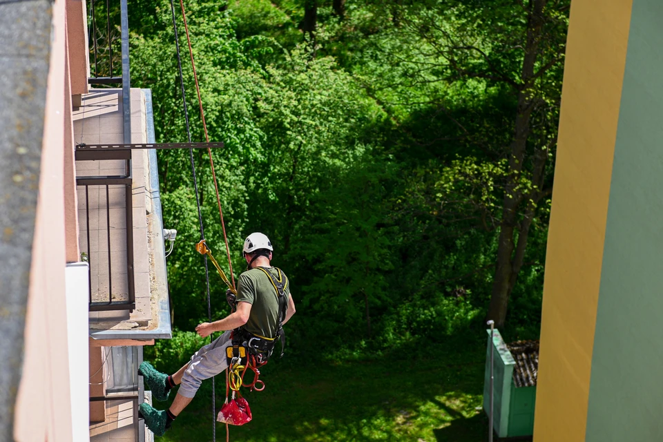 Cordiste équipé de harnais de sécurité, suspendu à une corde en façade de bâtiment, effectuant un test d'étanchéité avec pulvérisation d'eau colorée sur des joints de dilatation et menuiseries de fenêtres. Matériel de pulvérisation haute pression à la main, bâtiment urbain en arrière-plan, zone d'accès difficile en hauteur.